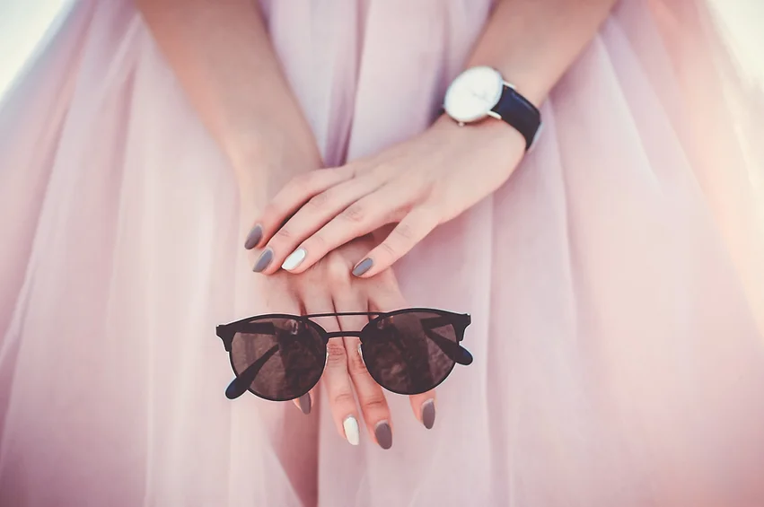 Partial image of a woman wearing a pink dress, clasping a pair of sunglasses with her hands.