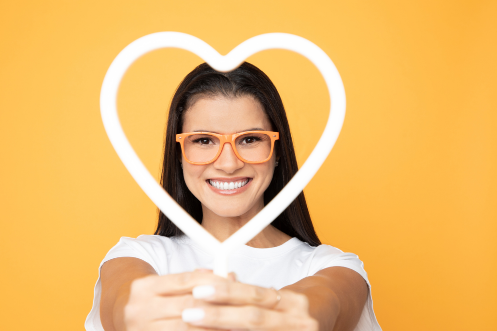 Woman wearing eyeglasses smiling and making a heart gesture against a bright yellow background.